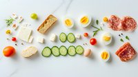 Fresh breakfast ingredients arranged in line on white background including bread, eggs, salami, cheese, vegetables and herbs for making sandwiches.