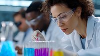 Mixed race female scientist in lab coat and safety glasses conducting research with colorful liquid samples in test tubes, focused on precise measurement.