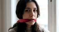 Young woman with red ribbon gag covering mouth in domestic setting expressing silence or censorship concept for awareness campaigns.