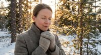 Peaceful young Caucasian woman with closed eyes enjoying warm winter sunlight in snowy forest wearing cozy gray coat and mittens during golden hour.