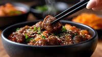 Steaming Asian meatballs in dark sauce garnished with sesame seeds and green onions served in black ceramic bowl, close up view with chopsticks.