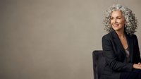 Mature woman with gray curly hair wearing black blazer, seated against neutral background, professional portrait for corporate and business use.
