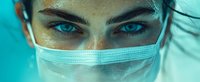 Close up portrait of young female medical worker with intense blue eyes and freckles wearing protective face mask against turquoise background.
