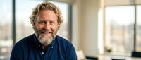 Middle-aged Caucasian businessman with curly hair and beard smiling confidently in modern office environment for corporate and professional content.