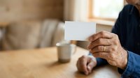 Senior man with dark skin holding blank white card at wooden table with coffee cup, suitable for business, greeting, announcement, or communication concepts.