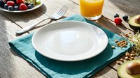 Empty white bowl on teal napkin with fresh fruit, orange juice and utensils arranged on wooden table for breakfast or brunch dining setup.
