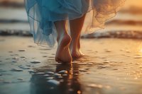 Barefoot woman in flowing dress walking on wet sand at sunset beach, with gentle waves reflecting golden light creating dreamy atmosphere.