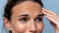 Mature Caucasian woman with wrinkled forehead showing signs of aging against blue background, expressing concern or worry with hand gesture.