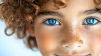 Close-up portrait of young person with bright blue eyes, natural freckles across nose and cheeks, and curly brown hair against light background.