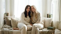 Two young women friends sitting together on beige sofa in bright living room, smiling and bonding. Cozy home interior with natural light for lifestyle content.
