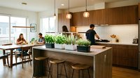 Warm, modern kitchen with wood cabinetry and island featuring potted herbs, pendant lighting, and family dining area with multiple generations enjoying meal together.