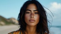 Young Asian woman with windswept dark hair against blurred coastal background, natural makeup and glowing skin, warm sunset lighting creates atmospheric portrait.