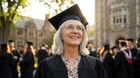 Mature Caucasian woman graduate in cap and gown smiling proudly at university ceremony with historic campus buildings in background.