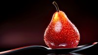Red pear covered in water droplets on black spoon against dark background, dramatic lighting creates glistening effect on wet fruit surface.