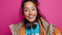 Young mixed race woman with bright smile wearing orange jacket and headphones against vibrant pink background expressing joy and positive emotions.