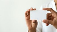 Young adult male with medium to dark skin tone wearing a white long-sleeved shirt, holding a blank white business card or paper between both hands at chest level against a neutral background.