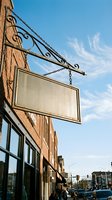 Vintage blank hanging sign with ornate iron bracket on brick building facade against blue sky for business storefront mockup and branding applications.