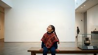 Young South Asian woman sitting on wooden bench in modern art gallery, wearing red plaid shirt, contemplating artwork on white wall, museum interior design.