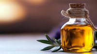 Small glass bottle of golden olive oil with cork stopper and twine, accompanied by fresh olive leaves on blurred background.