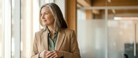Mature woman with long brown hair and fair complexion wearing a beige blazer and teal blouse, standing by a window in a modern office setting, looking thoughtful and professional.