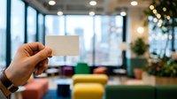 Hand holding blank white business card in modern office lounge with colorful seating and city skyline view. Mockup for corporate branding, marketing materials, and professional networking presentations.