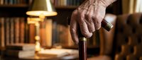 Senior man's weathered hand gripping wooden cane in home library with warm lighting, representing aging, mobility assistance, and indoor lifestyle for healthcare and senior living concepts.