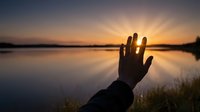 Hand silhouette reaching toward golden sunset over calm lake water with dramatic sky rays creating peaceful meditation moment for wellness concepts.