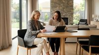 Two middle-aged women, one with blonde hair and one with dark hair, both of Caucasian ethnicity, sit at a wooden table reviewing documents and using a laptop together in a bright, modern home office with large windows and contemporary furnishings.