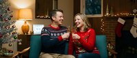 Cheerful Caucasian couple in holiday sweaters toasting with champagne on a cozy sofa near a decorated Christmas tree at home.
