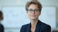 Professional middle aged Caucasian businesswoman with short blonde hair and glasses wearing navy blazer, confident smile in office setting with blurred background.