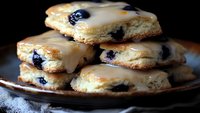 Flaky pastries filled with fresh blueberries and topped with sweet vanilla glaze stacked on rustic brown plate, close-up view showing layers of buttery dough.