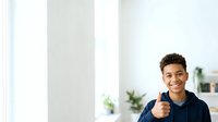 Young man with dark hair and medium skin tone wearing navy blue hoodie giving thumbs up gesture in bright modern interior with white walls and green plants, expressing approval and positivity.
