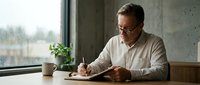 Middle-aged man with gray hair and glasses, wearing a beige shirt, writing at a desk by a window with a potted plant, natural daylight, professional workspace setting.