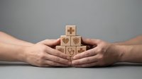 Two hands protectively cupping wooden blocks with healthcare symbols including hearts and medical cross on neutral background.