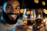 Young African American man with beard smiling while holding wine glass with sparkling bokeh lights in background, celebrating special occasion at night.