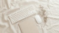 Minimalist home office flatlay with white keyboard, mouse, notebook and dried eucalyptus on soft beige linen fabric background.