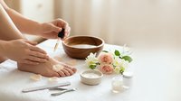 Woman applying nail polish to toenails during home pedicure with wooden bowl, roses and spa accessories on white towel for self-care and beauty routine.