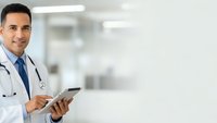 Confident Hispanic male doctor in white coat holding tablet computer in modern hospital corridor with copy space for medical technology concepts.