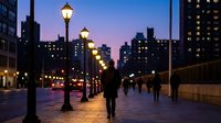 Urban evening scene with pedestrians walking along illuminated street lined with vintage lampposts against purple twilight sky and city buildings backdrop.