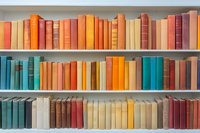 Vintage hardcover books arranged by color on white wooden shelves, creating rainbow gradient pattern from orange and red to turquoise and cream tones.
