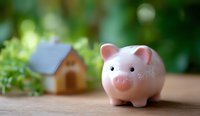 Pink ceramic piggy bank with small wooden house in blurred background, representing savings, investment and real estate planning concept on natural bokeh.