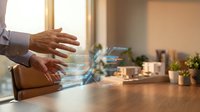 Businessman with brown skin tone gesturing over wooden desk with holographic digital interface and data visualization, representing technology innovation and business solutions in modern office environment.