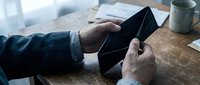 Senior businessman hands holding empty black leather wallet at wooden desk with coffee cup and papers in background showing financial difficulty concept.