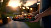 Grilled meat and vegetables served on black plate during sunset backyard dinner party with bokeh lights creating warm atmospheric ambiance.