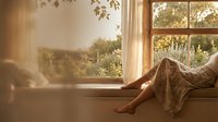 Woman relaxing by sunny window with warm golden light streaming through glass panes creating peaceful morning atmosphere in cozy home interior.