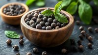 Aromatic black peppercorns in wooden bowl with fresh basil leaves on dark stone background, scattered spices and herbs for cooking and seasoning.