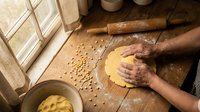 Elderly hands kneading fresh pasta dough on wooden surface with rolling pin and flour scattered around in rustic kitchen setting for cooking preparation.