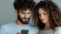 Young diverse couple looking at smartphone screen together, smiling and browsing social media. Mediterranean man with beard and curly-haired woman sharing digital content.
