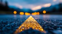 Glowing yellow road marking on wet asphalt at dusk creates leading line with bokeh lights in background, moody atmospheric evening scene with shallow depth of field.