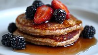 Stack of golden pancakes drizzled with maple syrup and topped with fresh strawberries and blackberries on white plate, close up view of breakfast food.